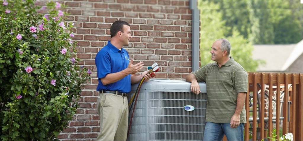 HVAC technician wearing blue shirt talking to homeowner next to outdoor HVAC unit.
