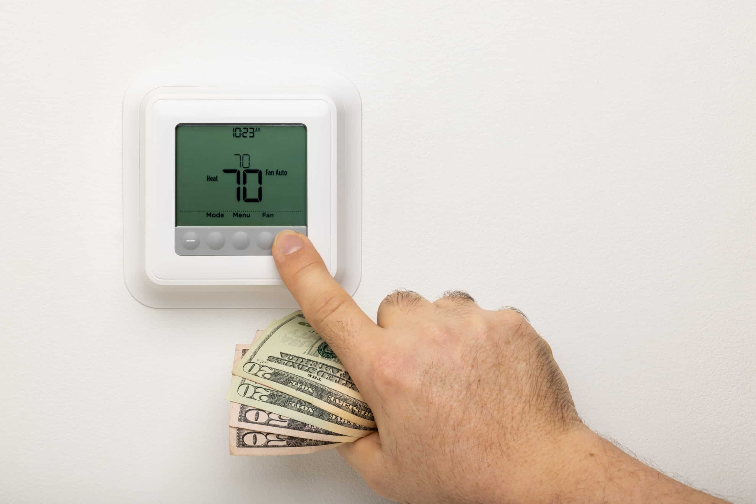 A hand holding money while also adjusting a thermostat on a white wall with temperature reading of 70 degrees.